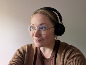 A woman with a slight smile and gold-rimmed glasses with noise cancelling headphones on wearing a brown sweater.