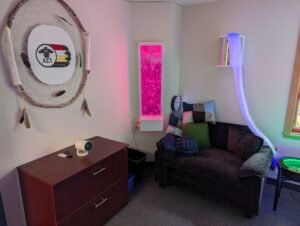 A large dreamcatcher with AEI's logo on it hanging in Nipissing Campus's Sensory Lounge. There is a mounted bubble rube on the wall, an oversized chair with sensory pillows, and other sensory lighting next to a window.