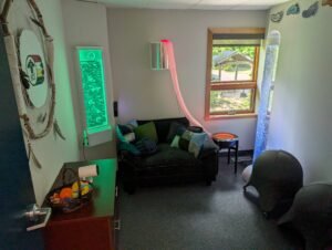 A large dreamcatcher with AEI's logo on it hanging in Nipissing Campus's Sensory Lounge. There is a mounted bubble rube on the wall, an oversized chair with sensory pillows, and other sensory lighting next to a window with a stand up bubble tube and two ball chairs.